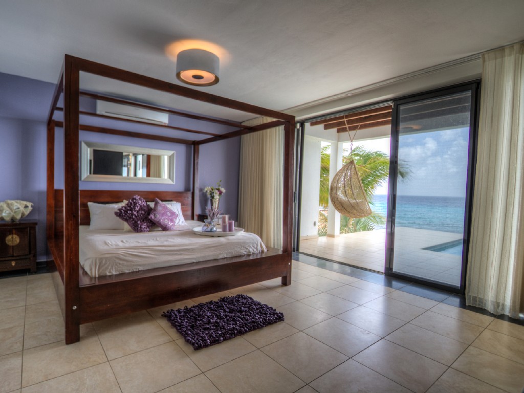 Luxury Bonaire villa master suite with a dark wood four-poster bed, private terrace with hanging chair, and stunning direct ocean and plunge pool views.