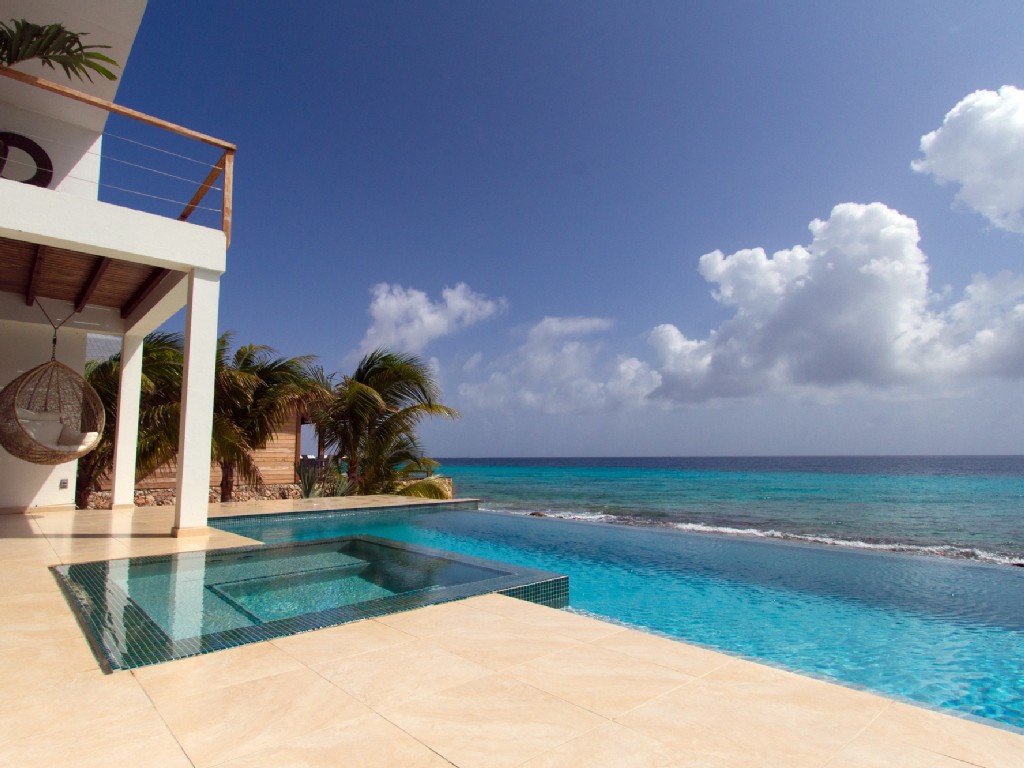 Luxury Bonaire villa with an oceanfront infinity pool, modern travertine terrace, hanging wicker chair, and panoramic views of the turquoise Caribbean Sea under a blue sky.
