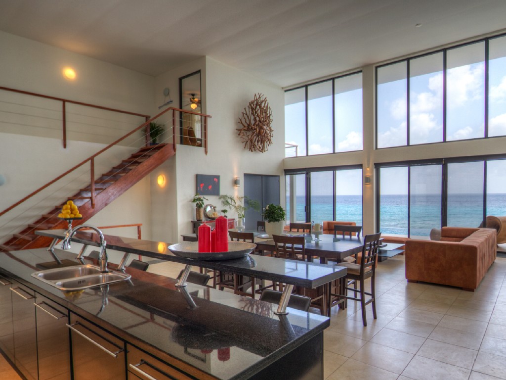 Luxury Bonaire villa great room with double-height windows, panoramic ocean views, a granite kitchen island, and modern open-riser staircase.