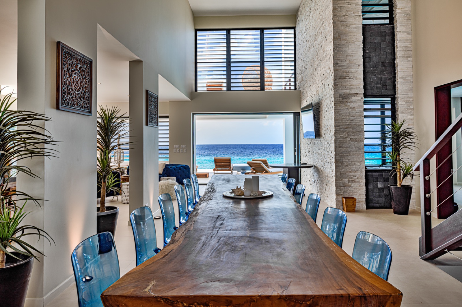 Luxury Bonaire villa dining room with a long live-edge wood table and modern blue chairs, opening to a stunning ocean view terrace and pool.