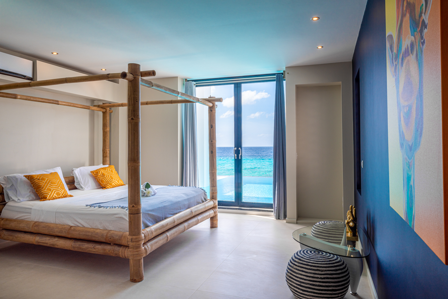 Luxury Bonaire villa master suite with a bamboo canopy bed and floor-to-ceiling glass doors opening onto an infinity pool with a panoramic Caribbean Sea view.