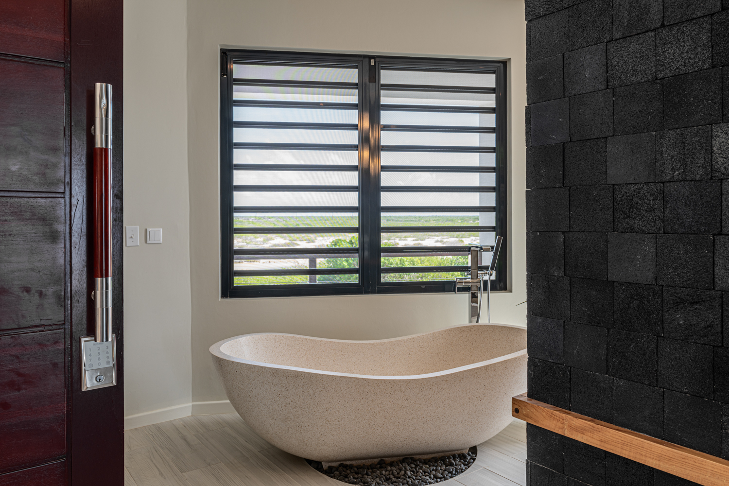 Luxury Bonaire villa spa bathroom featuring a freestanding terrazzo soaking tub on a bed of river rocks, a dramatic black lava stone wall, and a view of the natural landscape.