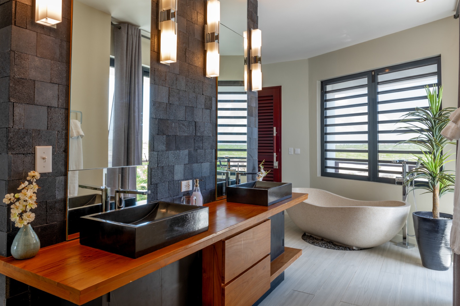 Luxury Bonaire villa spa bathroom with a freestanding stone soaking tub, double vanity with black vessel sinks, and a dramatic black lava stone accent wall.