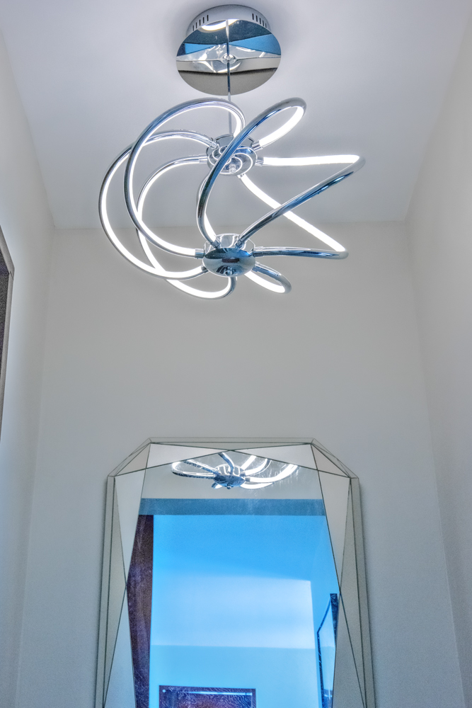Modern sculptural light fixture with chrome finish in a luxury Bonaire villa, reflected in a large beveled geometric wall mirror against a white wall.
