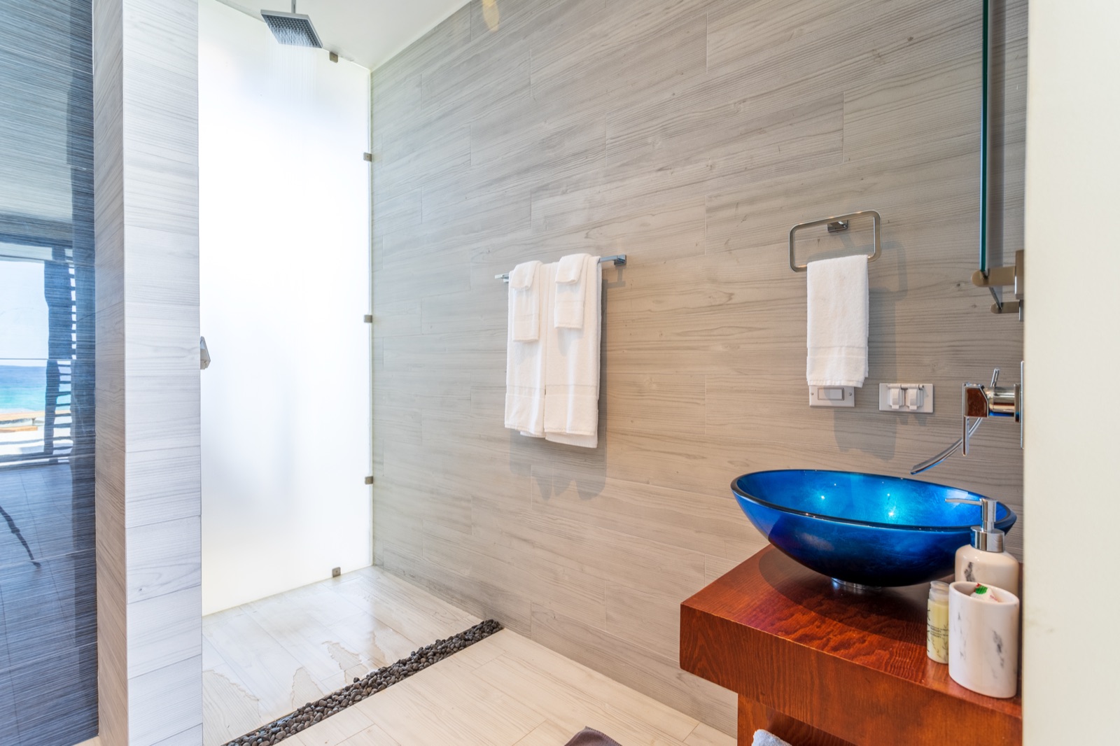 Luxury Bonaire villa spa bathroom with a walk-in rain shower, cobalt blue glass vessel sink on a wood vanity, and a reflection of the ocean view.