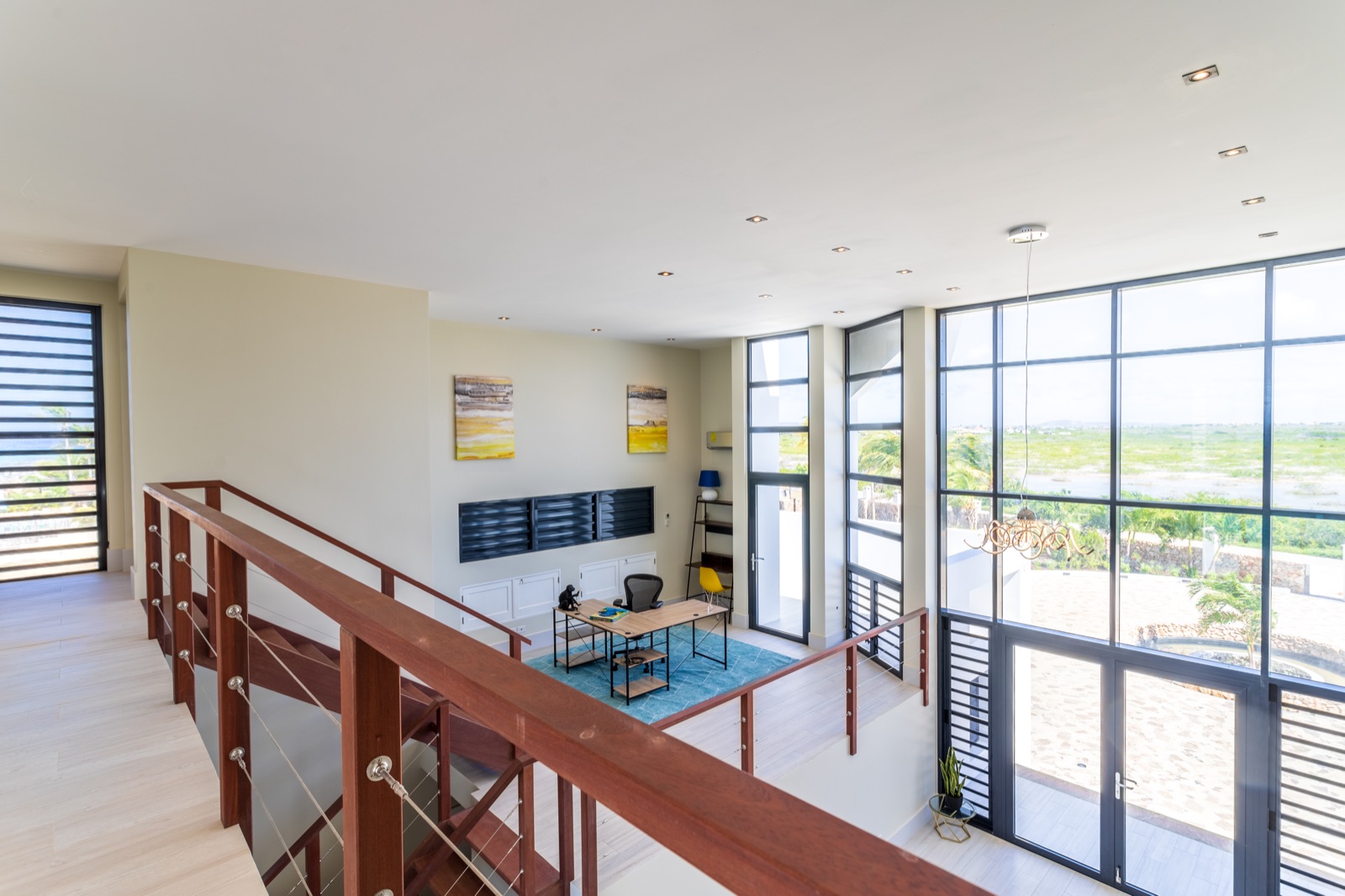 Luxury Bonaire villa mezzanine office with double-height ceilings, modern wood staircase, and floor-to-ceiling windows with panoramic nature views.