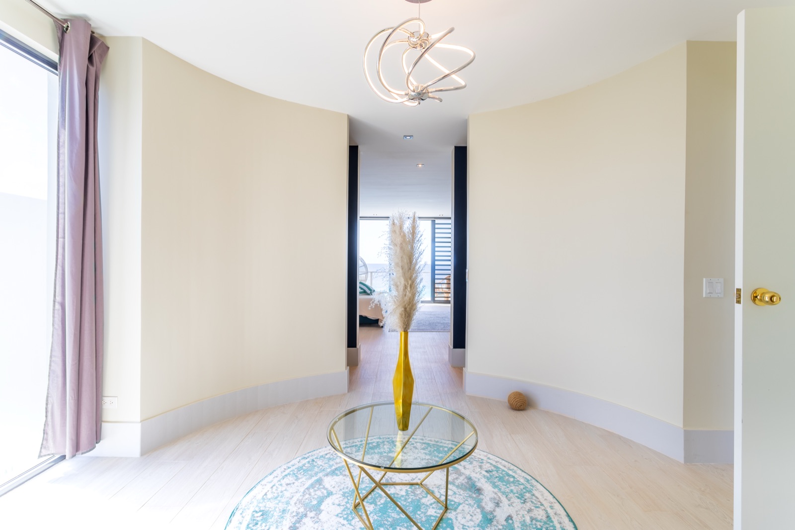 Curved entryway to a luxury Bonaire villa master suite, featuring light wood floors, a modern sculptural chandelier, and a view into the sunlit bedroom.