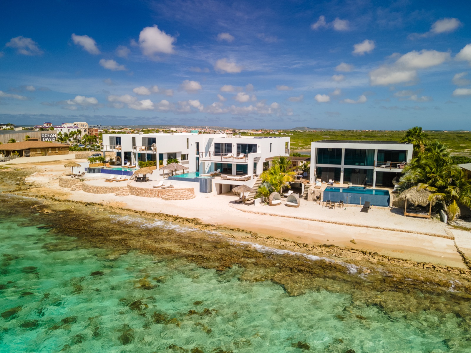 Aerial view of luxury Bonaire villas with modern white architecture, private infinity pools, and direct beachfront access to the turquoise Caribbean Sea under a blue sky.